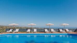 Piscina com cadeiras de apoio brancas e guarda-sóis, céu claro e vista distante do mar e montanhas.