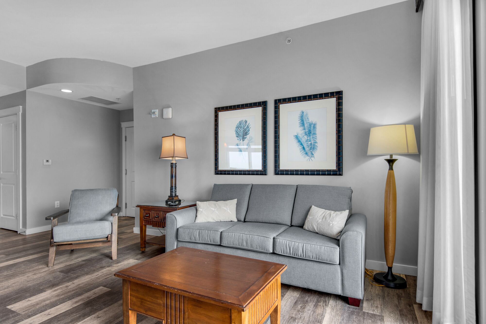 A modern living room with a grey sofa, a wooden coffee table, a side table with a lamp, and framed artwork on the wall.