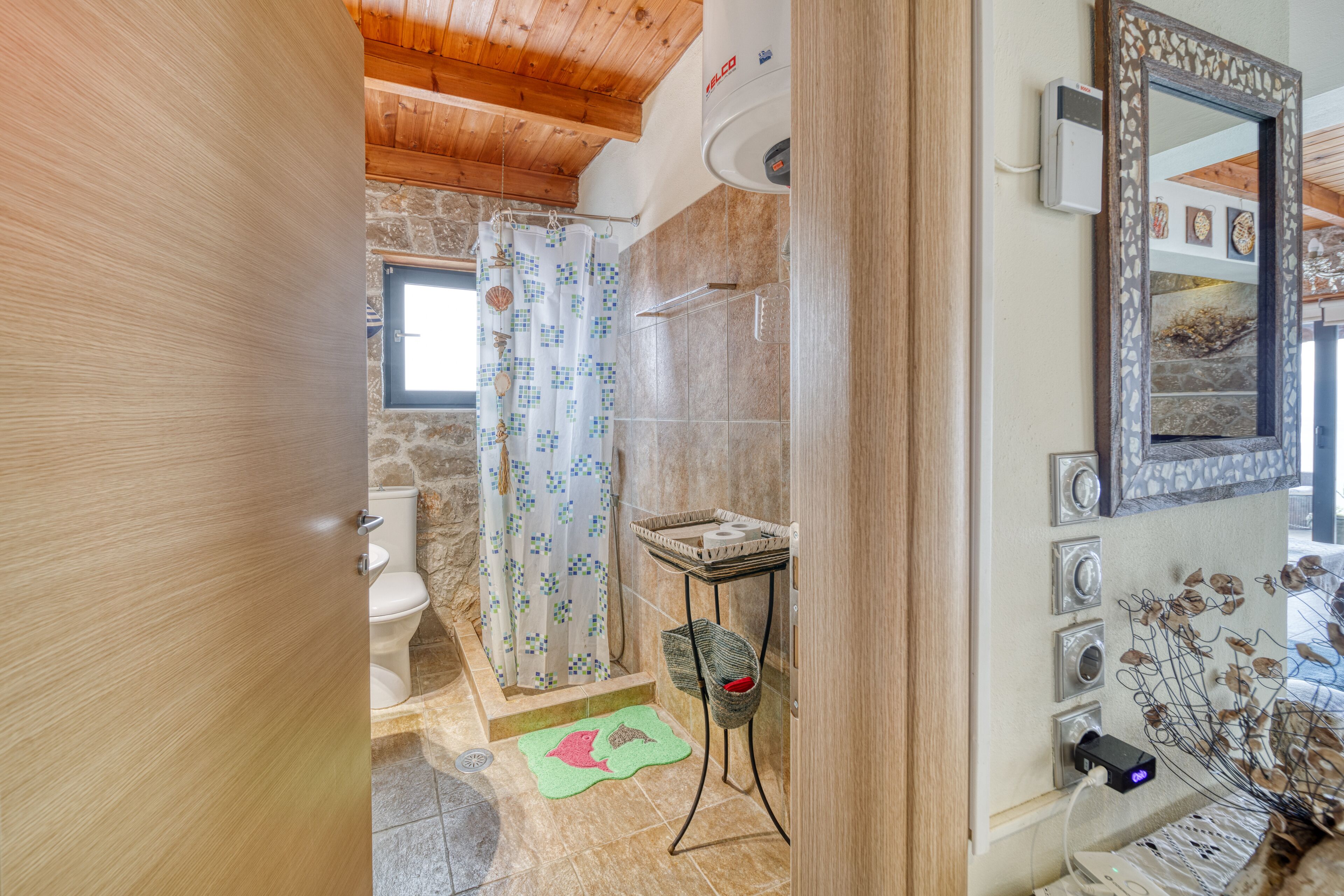 Ein Badezimmer mit Steinwand, Holzboden, Dusche, Toilette, Waschbecken mit Korb und Spiegel.