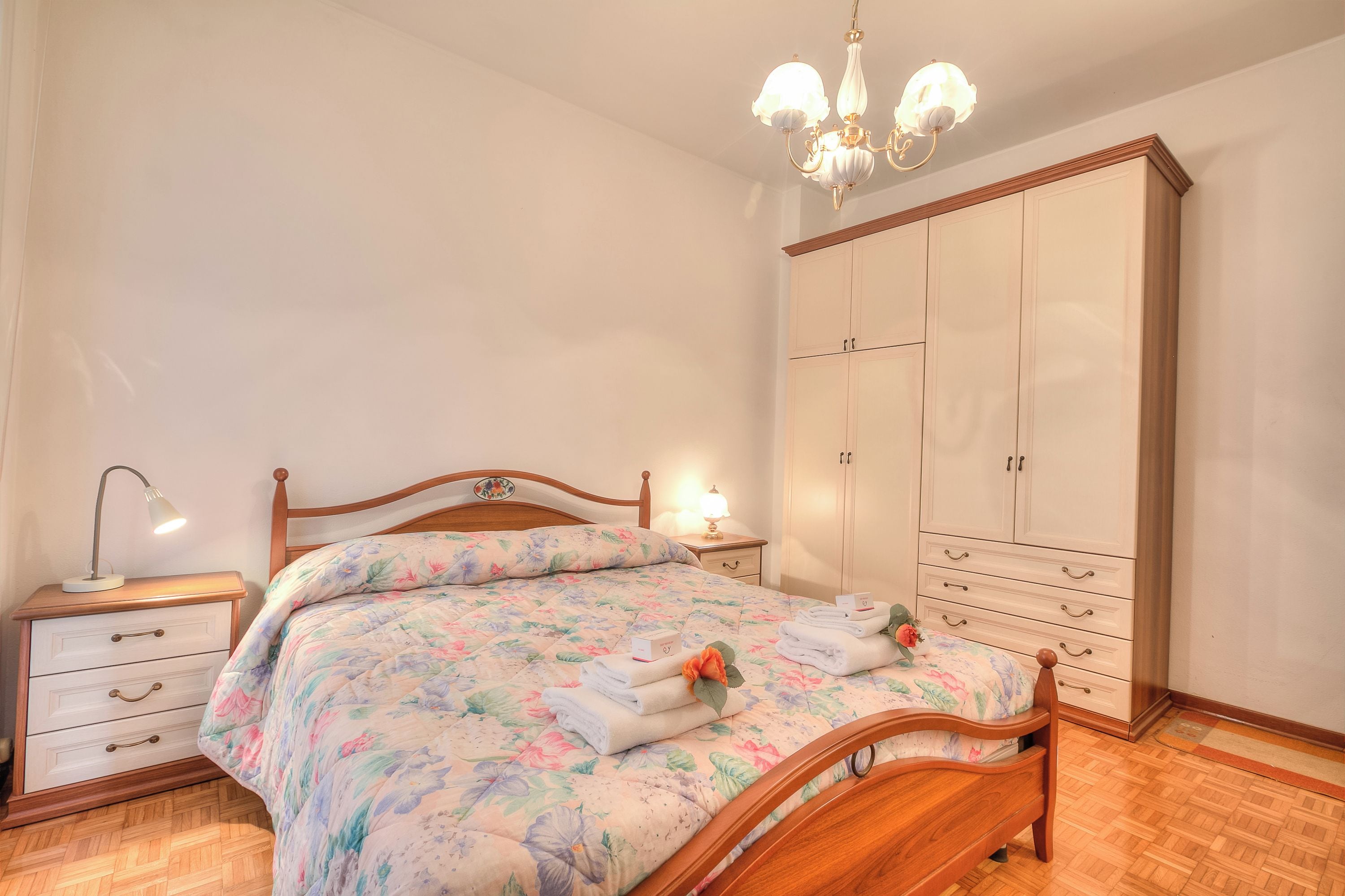 A bedroom with a wooden bed, a nightstand with a lamp, a wardrobe, and a chandelier.