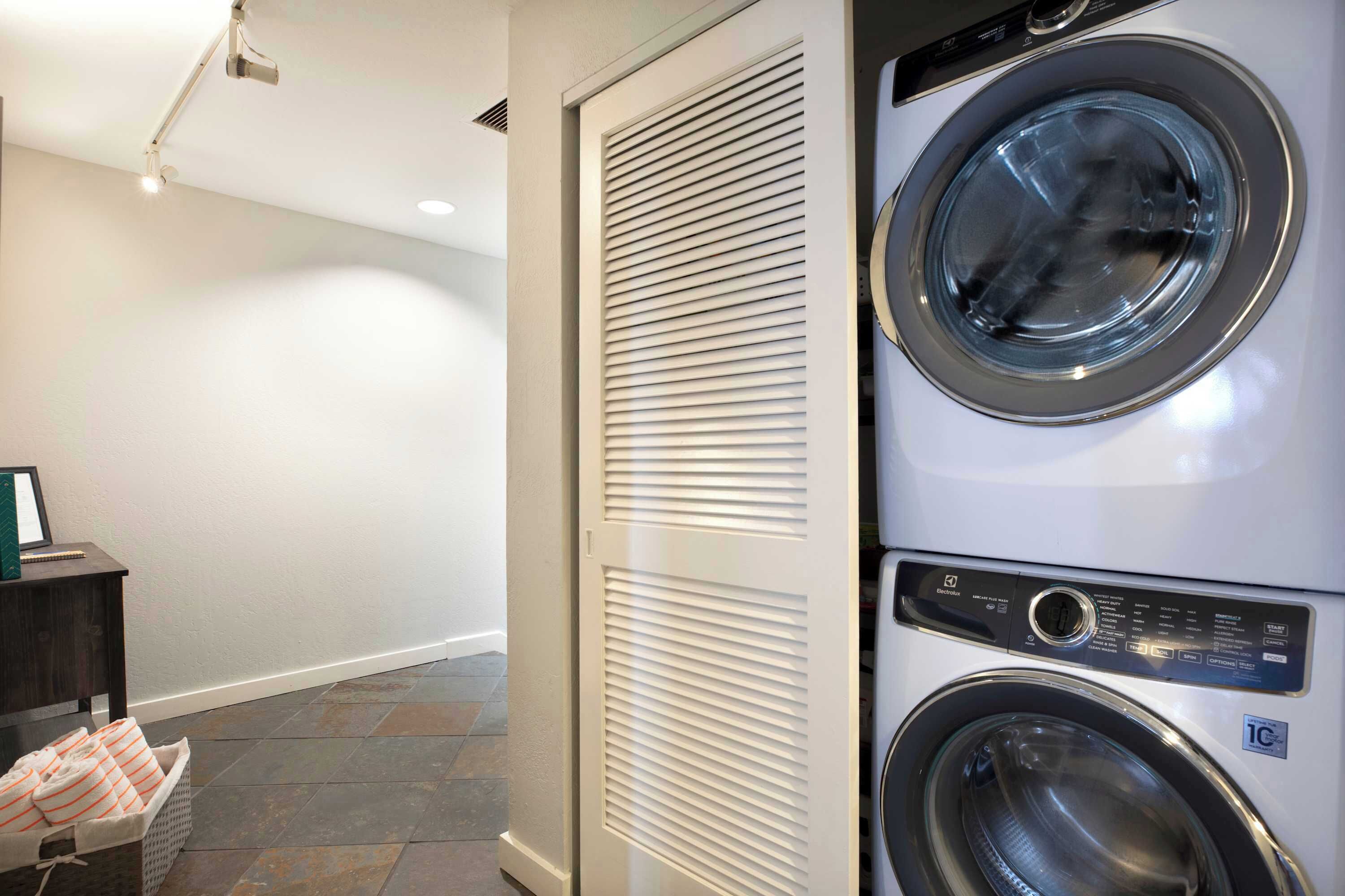 A laundry room with a stacked washer and dryer, a small table with a basket of folded towels, and a wall-mounted vent.