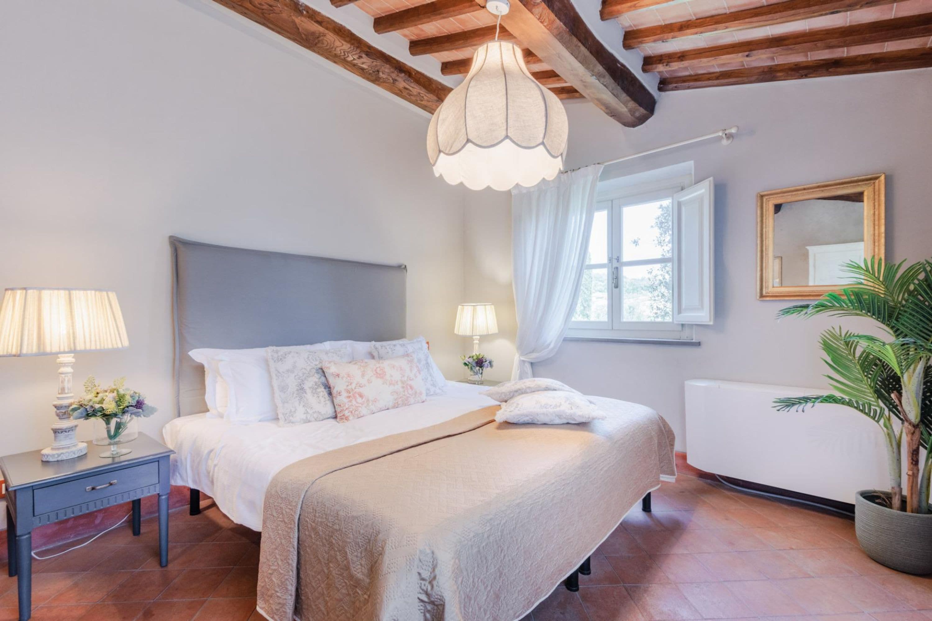 A bedroom with a bed, bedside lamps, a window with curtains, a radiator, and a potted plant.