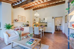 A spacious living area with a wooden coffee table, a white sofa, and a dining table with chairs. A kitchen with white cabinets and a tiled backsplash is visible in the background.