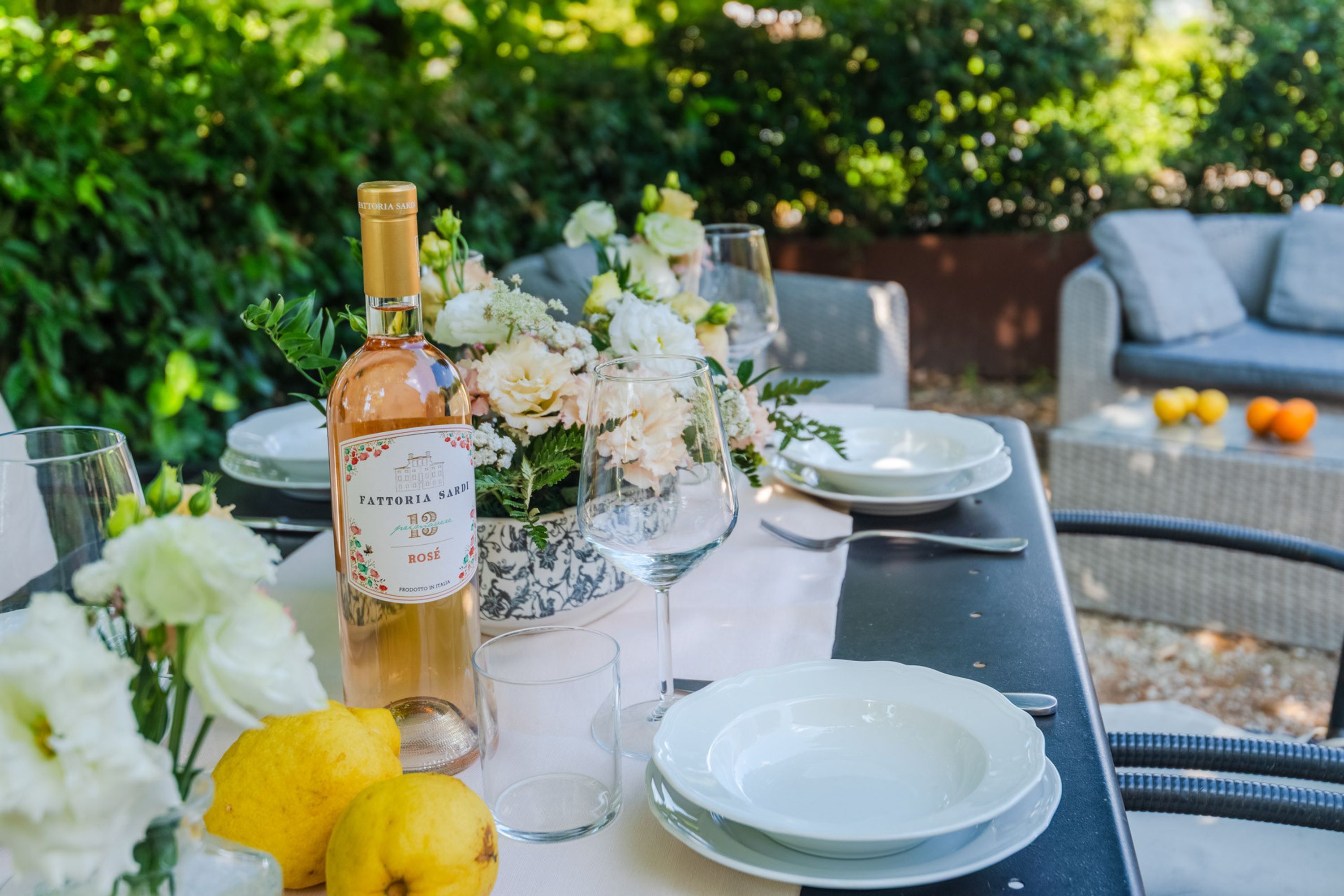 An outdoor dining table set with a bottle of Fattoria Sardi wine, glasses, plates, and a centerpiece of flowers.