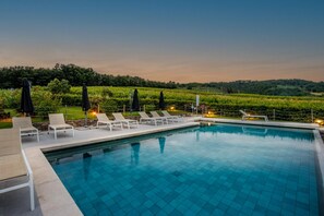 A swimming pool with tiled flooring, surrounded by lounge chairs and umbrellas, set against a backdrop of a lush green landscape.