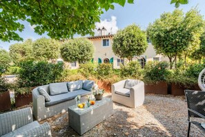 An outdoor seating area with wicker furniture, a glass coffee table with fruits and flowers, and a view of a building with a tiled roof and greenery.