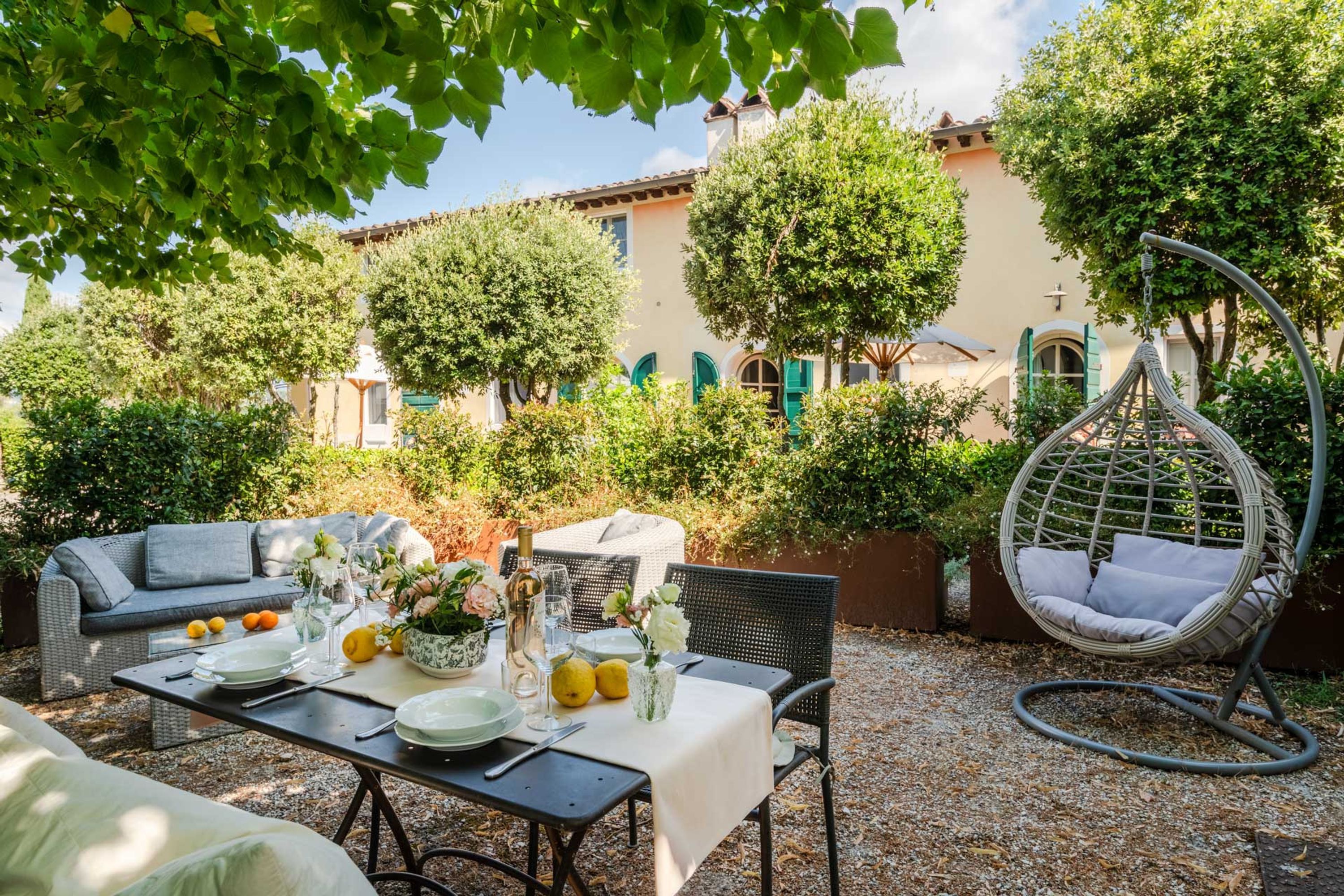 An outdoor dining area with a table set for four, surrounded by greenery and a hanging chair.