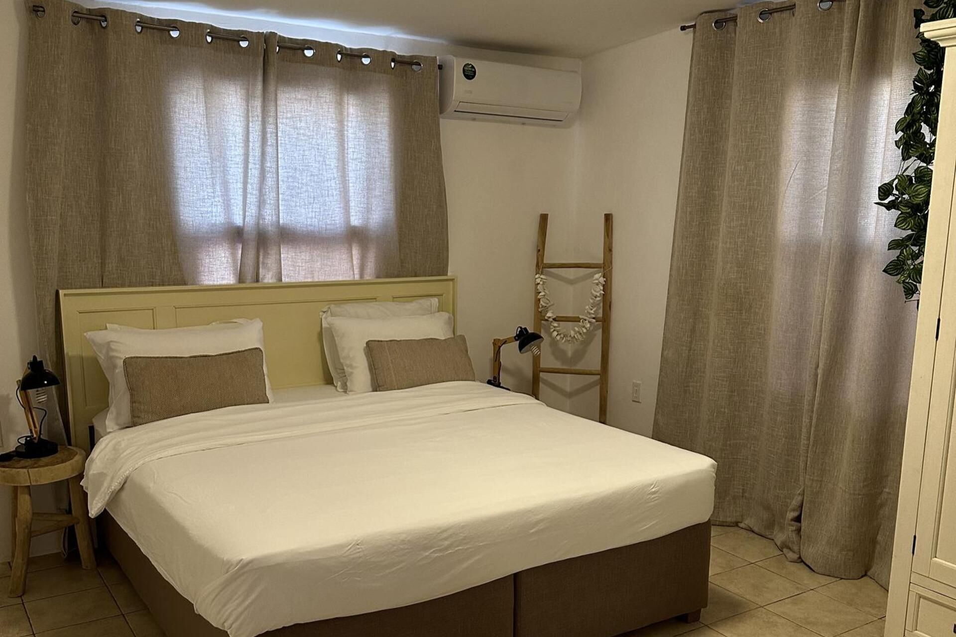 A bedroom with a bed, curtains, an air conditioning unit, a ladder, and a small table with a lamp.