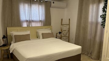 A bedroom with a bed, curtains, an air conditioning unit, a ladder, and a small table with a lamp.