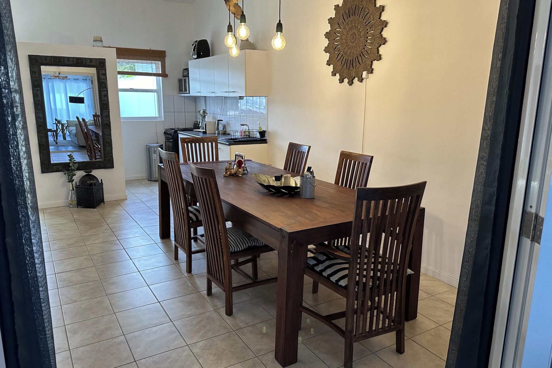 A dining area with a wooden table and chairs, a kitchen in the background, and a decorative wall piece.