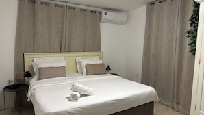 Una cama bien hecha con sábanas blancas y una toalla enrollada, una mesita de noche con lámpara, y un aire acondicionado instalado en la pared.