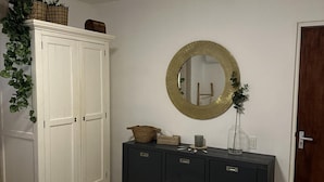 Una habitación con un armario blanco, un aparador oscuro con cajones, un espejo redondo con marco dorado y una puerta de marco de madera.