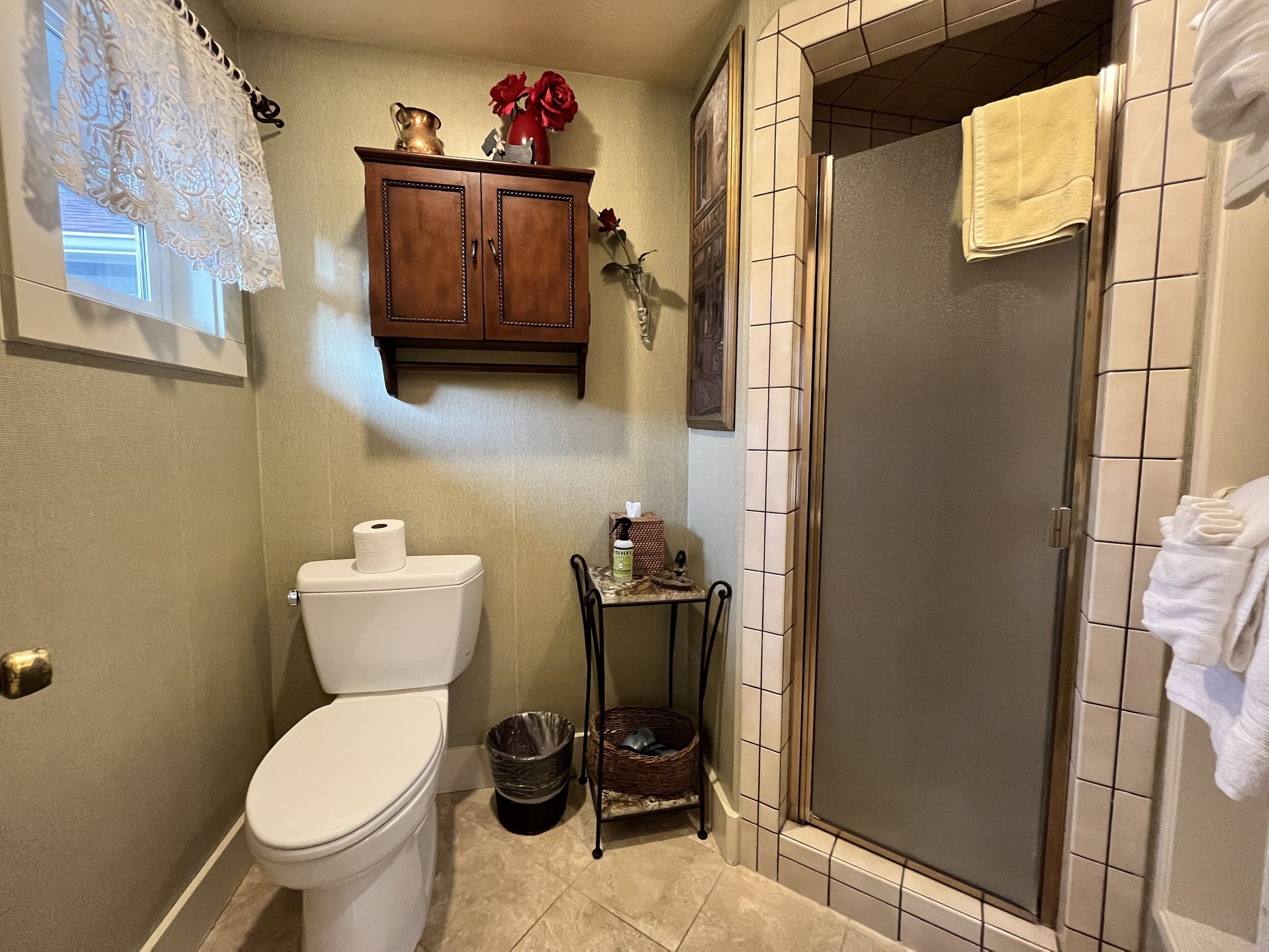 A bathroom with a toilet, a trash can, a small table with a basket, and a shower area with a towel hanging on the door.