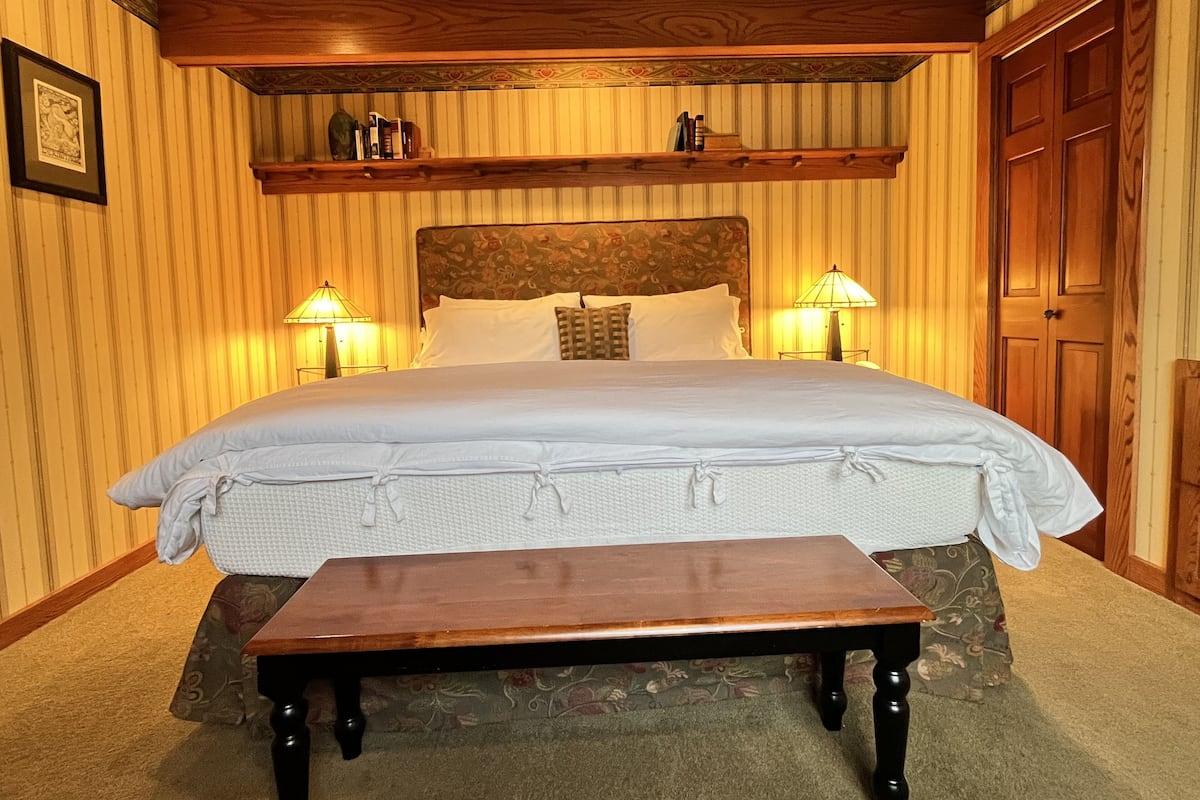 Un dormitorio ordenado con una cama grande, dos lámparas de noche, un cabecero de madera, una mesita y una puerta.