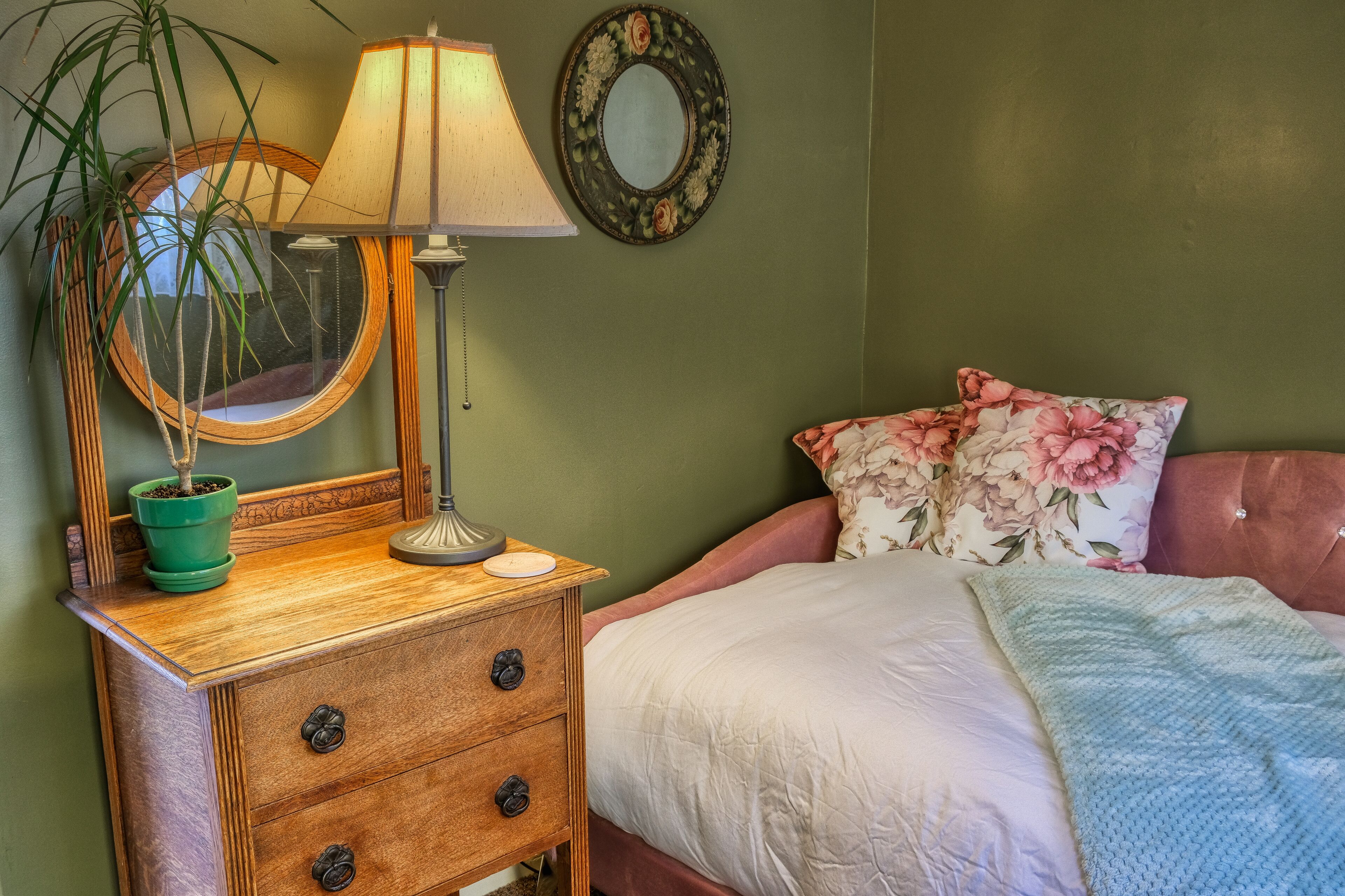 Een slaapkamer met een houten nachtkastje, een lamp, een spiegel, een groene plant in pot, een bloemenkussen en een bed met een wit- en blauwe deken.
