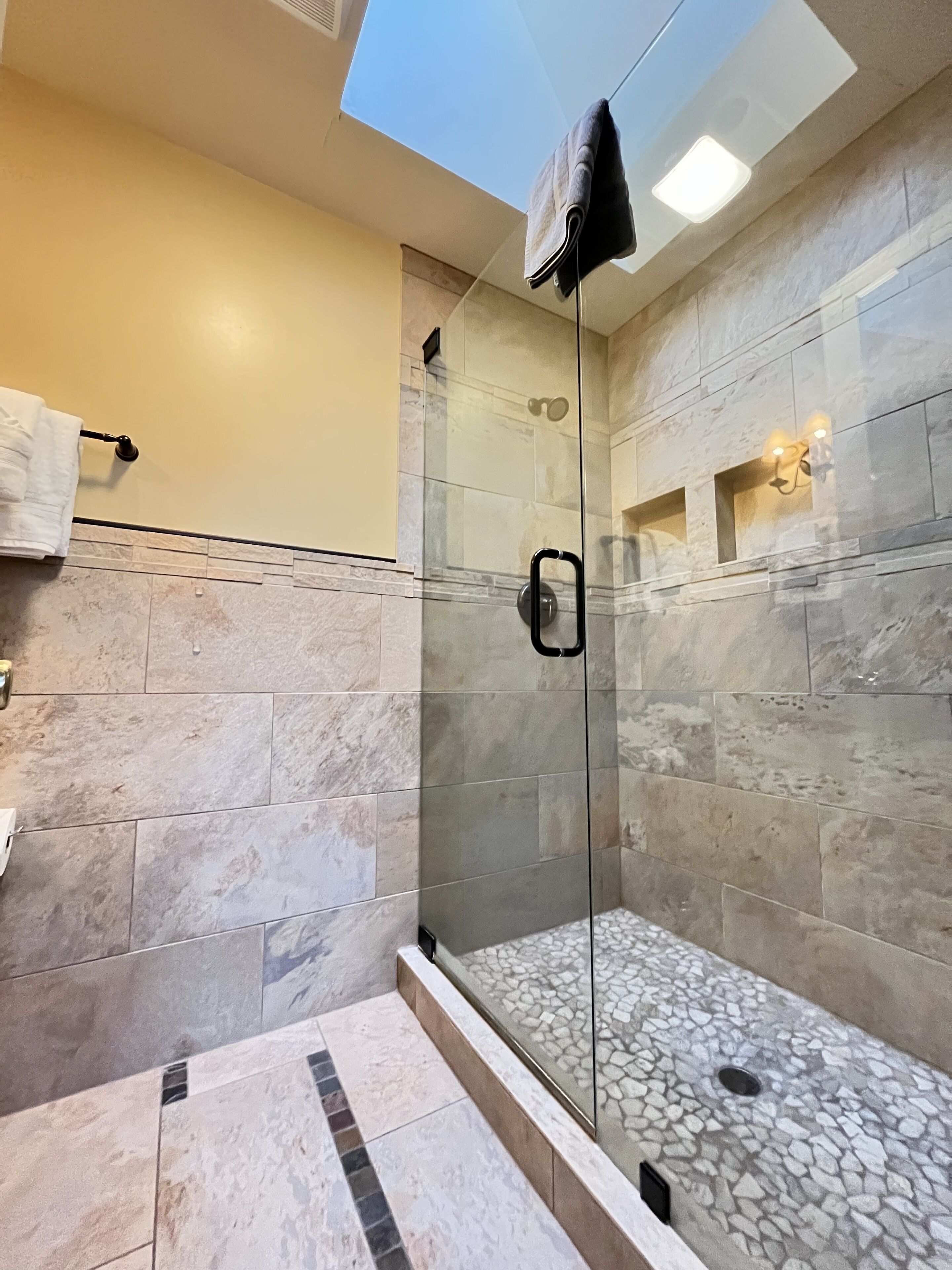 A modern bathroom with a glass-enclosed shower, marble tiles, and a skylight.