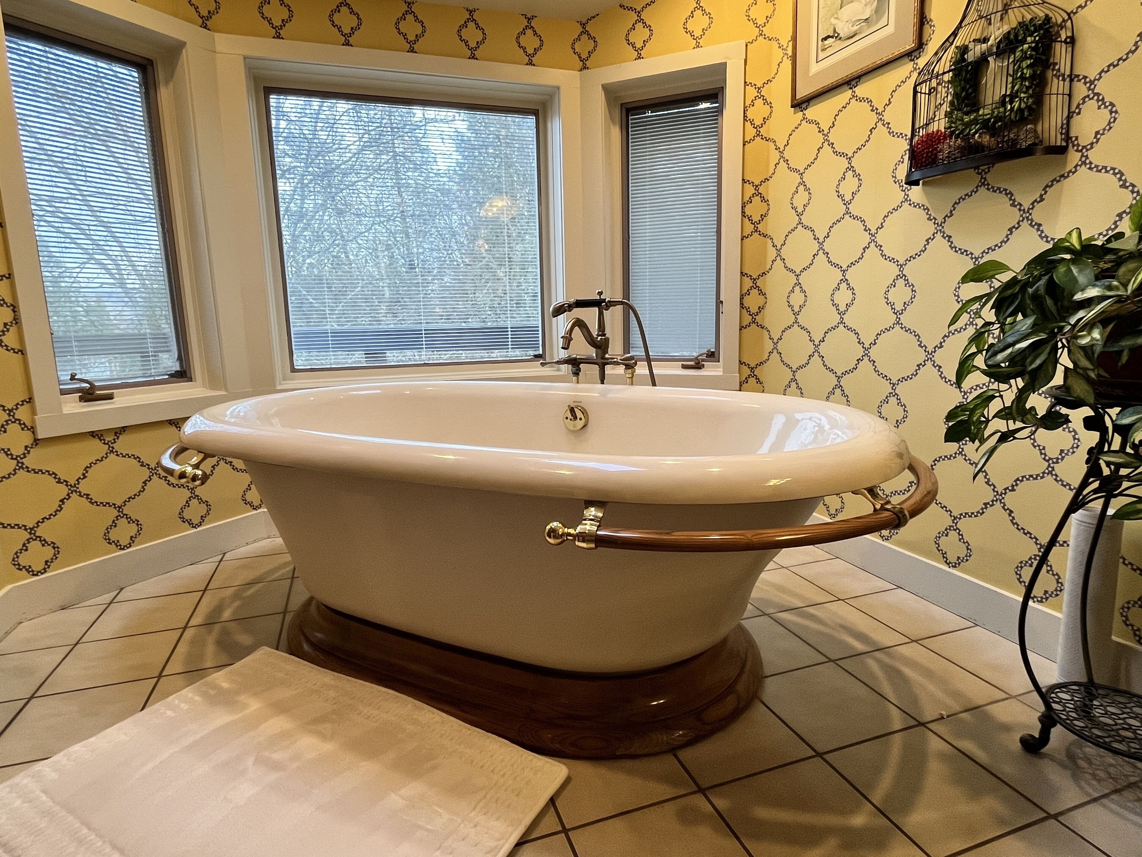 A bathroom with a freestanding bathtub, two windows, patterned wallpaper, and a potted plant.