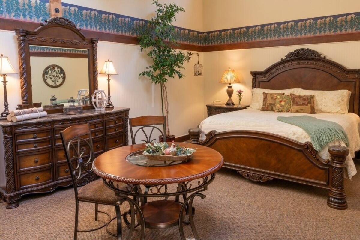 Un dormitorio con una cama de madera, un aparador, una mesa redonda con sillas, un espejo y una planta en maceta.