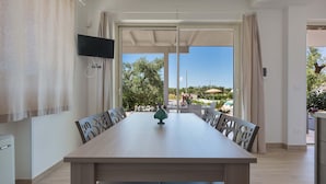 Un espace repas comprenant une longue table, des chaises, un téléviseur fixé au mur et une vue sur l’extérieur.