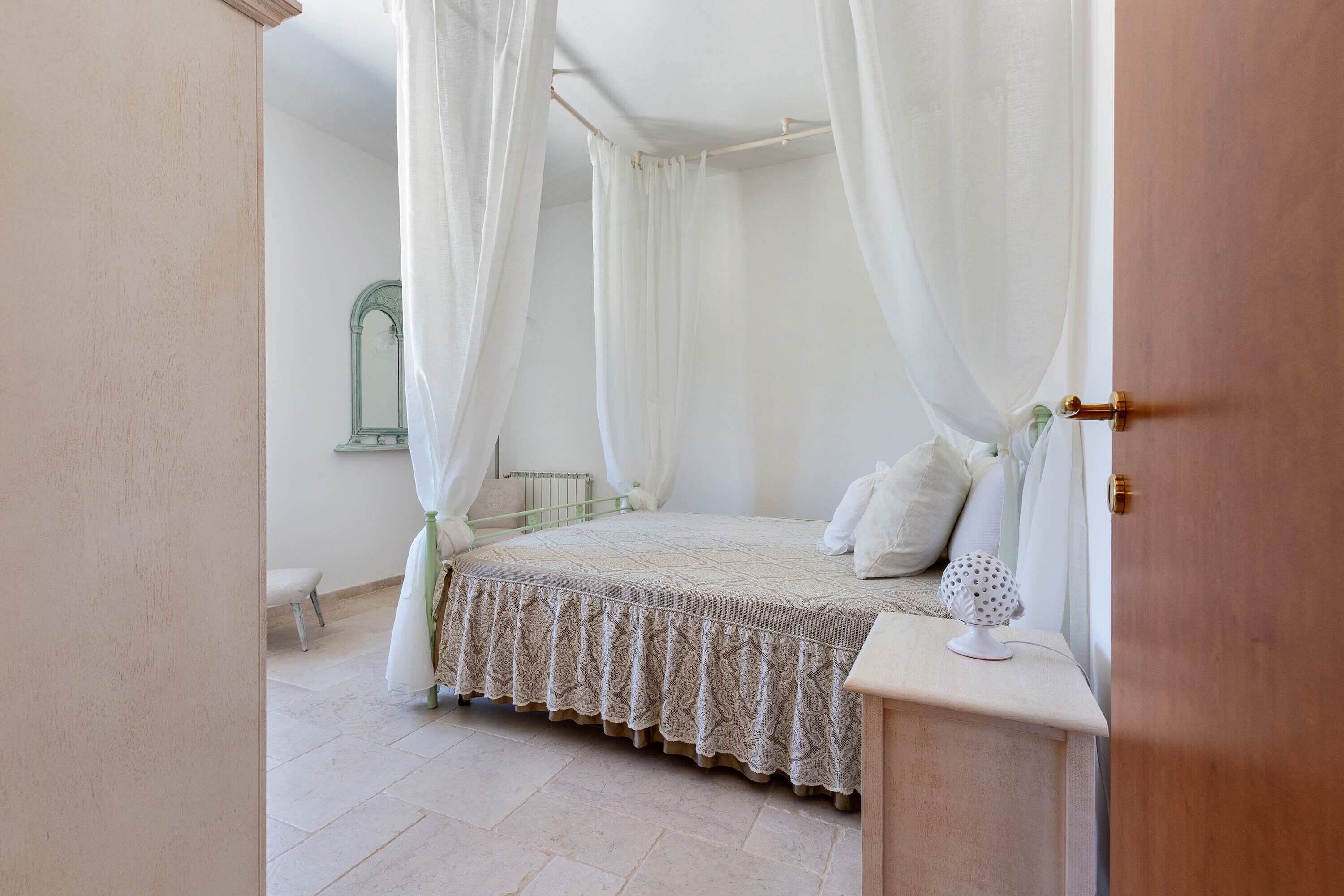 Un dormitorio con cama con dosel, cortinas blancas y una mesita de noche con lámpara.