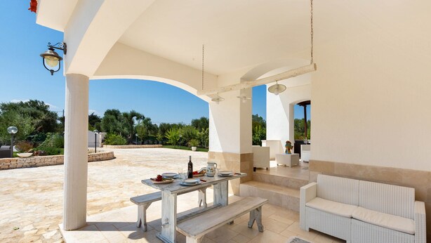 Une terrasse extérieure avec un canapé en osier blanc, une table en bois avec un banc et un luminaire suspendu.