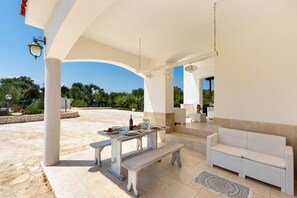 An outdoor patio with a white wicker sofa, a wooden table with a bench, and a hanging light fixture.