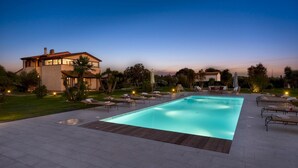 Una piscina rectangular con sillones reclinables y sombrillas, rodeada por un césped cuidado y una casa grande al fondo.