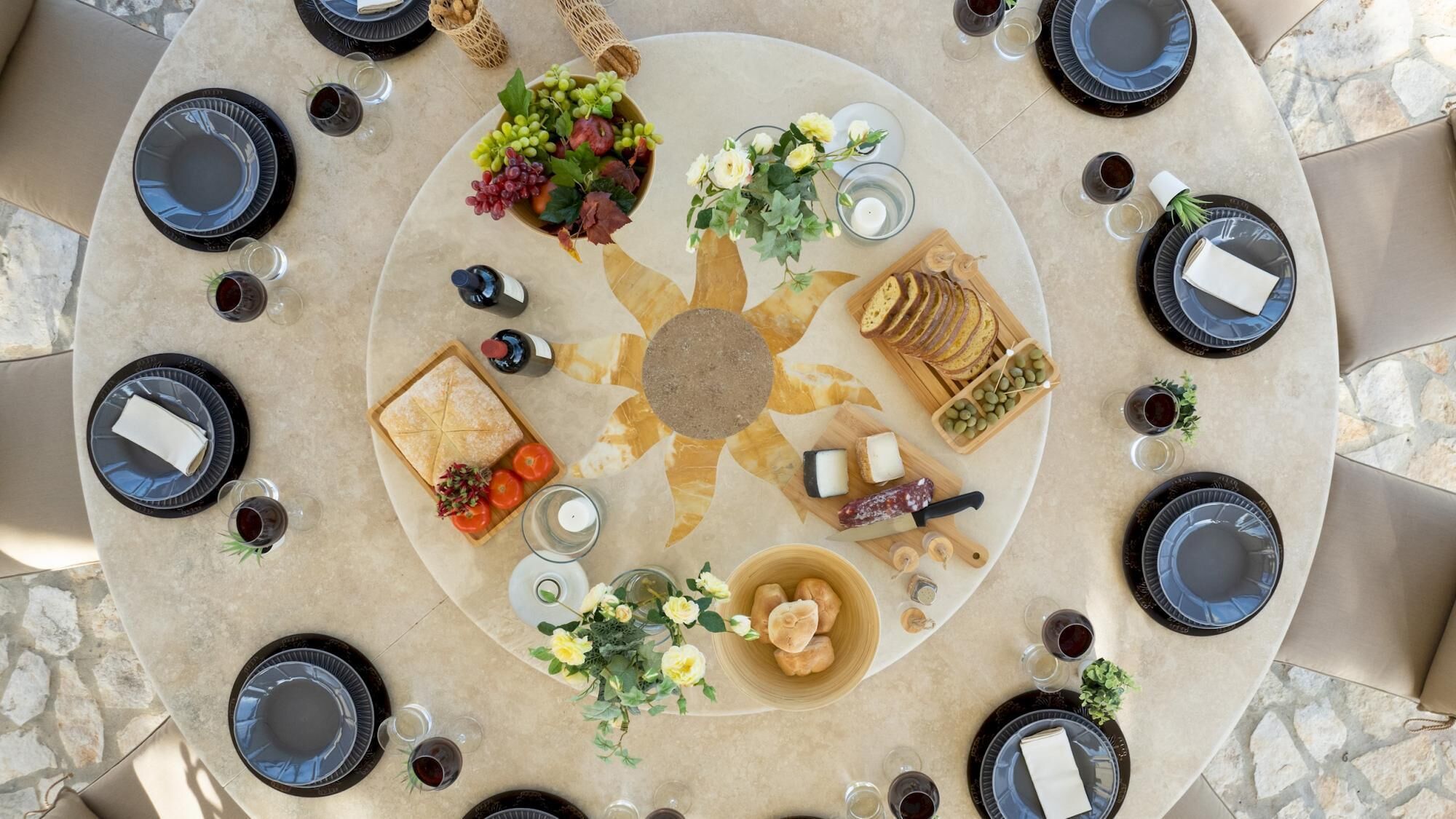 Una mesa redonda con un arreglo floral central, rodeada de platos, vasos y diversos alimentos.