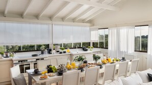 Una cocina luminosa con armarios blancos, un comedor con sillas blancas y ventanas amplias con vistas a zonas verdes.