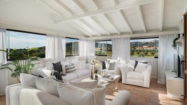 Un salón espacioso con muebles blancos, ventanas amplias que ofrecen vistas al campo, y un techo con vigas a la vista.