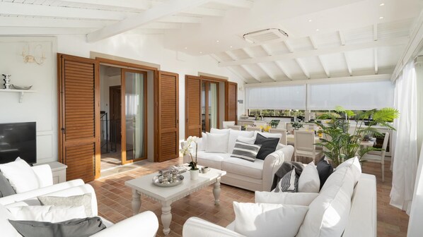 A spacious living area with a white sofa set, a coffee table, and a TV. There are wooden doors leading to a balcony with a view of the outdoors. - La Buganvilla 14 (Campiglia Marittima)