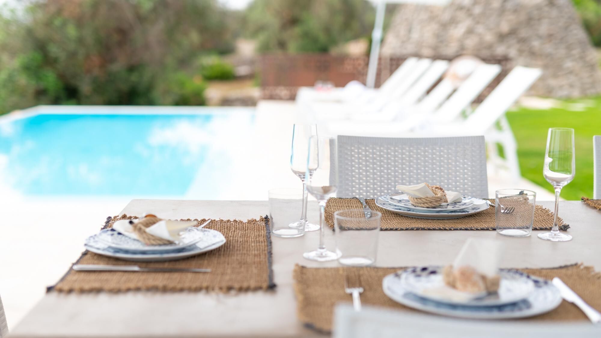 Una mesa de comedor dispuesta con platos, cubiertos y vasos, con vista a una piscina.