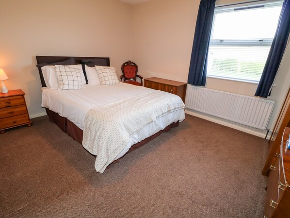 A neatly arranged bedroom with a bed, bedside table, lamp, chair, and window with curtains. - 17 Buninver Road (Omagh)