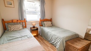 Un dormitorio con dos camas individuales, una mesita de noche con lámpara, un cofre de mimbre y una ventana con cortinas.