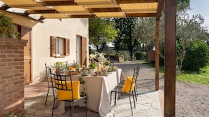 An outdoor dining area with a table set for two, surrounded by chairs with yellow cushions, under a wooden pergola. - Casa La Fenice 8 2 (Pomarance)