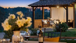 An outdoor dining area with a wooden pergola, tables, chairs, and a view of the mountains at dusk. - Casa La Fenice 8 2 (Pomarance)