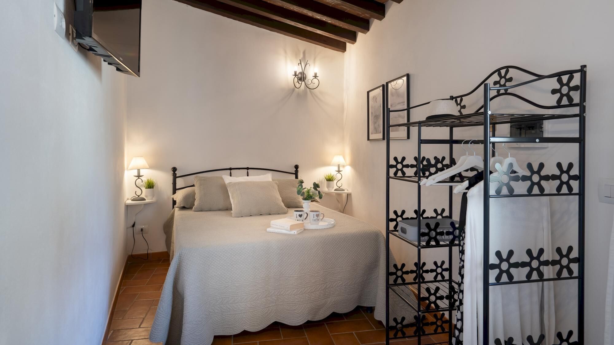 Un dormitorio ordenado con cama, lámparas de noche, una lámpara de pared, un perchero con ropa colgada y suelo de baldosas.