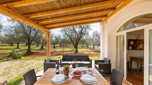 An outdoor dining area with a wooden table set for two, surrounded by wicker chairs, overlooking a grassy landscape with trees. - 2770 Uliveto Valeria - Trullo Melograno by Perle di Puglia (San Michele Salentino)