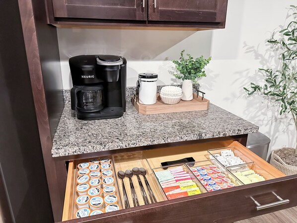 Un comptoir de cuisine avec une machine à café Keurig, une cafetière blanche et un plateau contenant des bols et une plante. En dessous, un tiroir renferme des dosettes de café et des ustensiles.