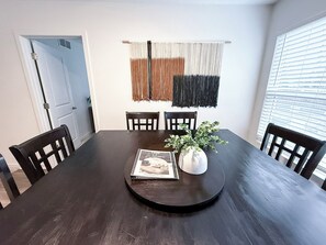 Une salle à manger avec une table en bois foncé, quatre chaises, un vase blanc contenant une plante et un magazine posé sur la table.