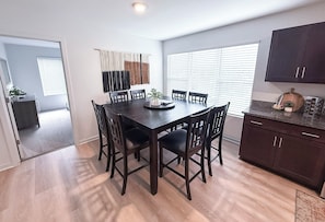 A dining area with a dark wooden table and chairs, a small plant on the table, and a kitchen area with dark cabinets and a countertop.