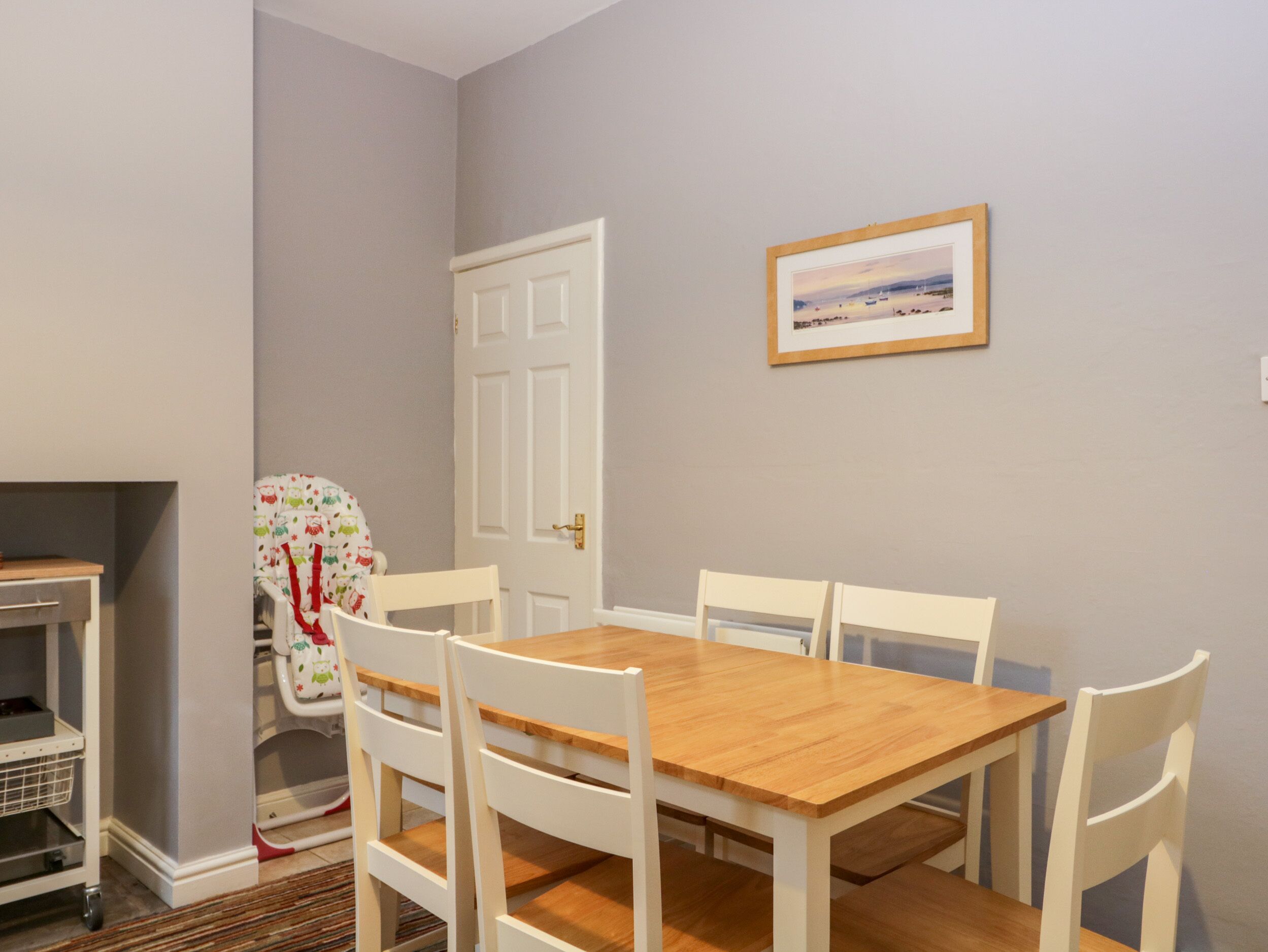 Un comedor con una mesa de madera y sillas blancas, una puerta blanca y un cuadro en la pared.