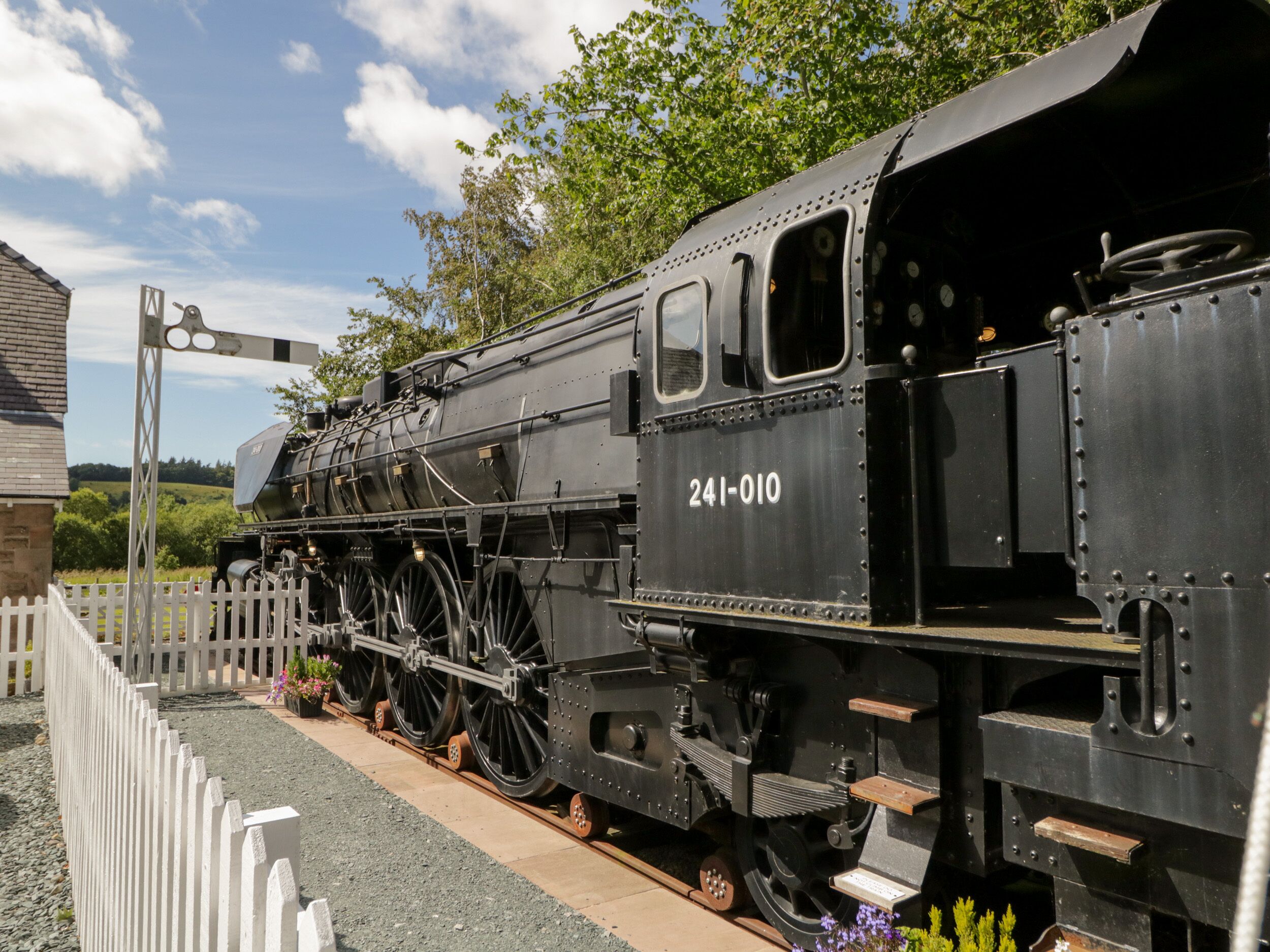 A black steam locomotive numbered 241-010 parked at a station with a white fence and greenery in the background.