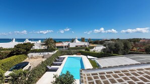 A rooftop swimming pool with a view of the sea, surrounded by greenery and modern buildings.