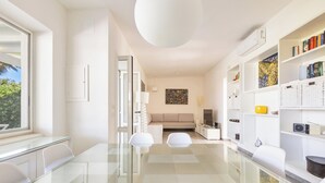 A modern living room with a glass dining table, white sofas, a beige sofa, and a bookshelf filled with books and decorative items.