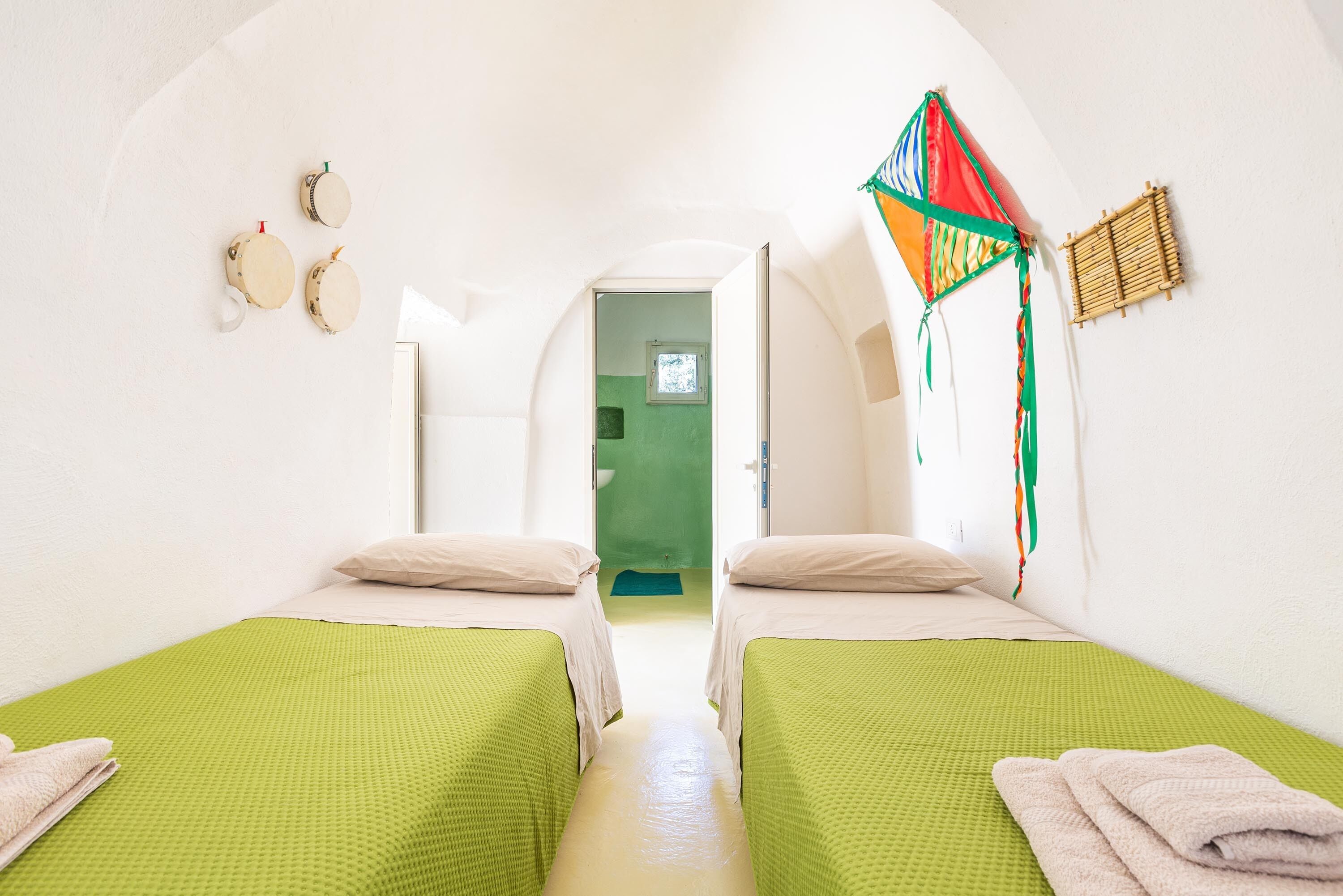 Una habitación con dos camas individuales, un suelo verde con textura, una puerta blanca que da al baño y una cometa de colores en la pared.
