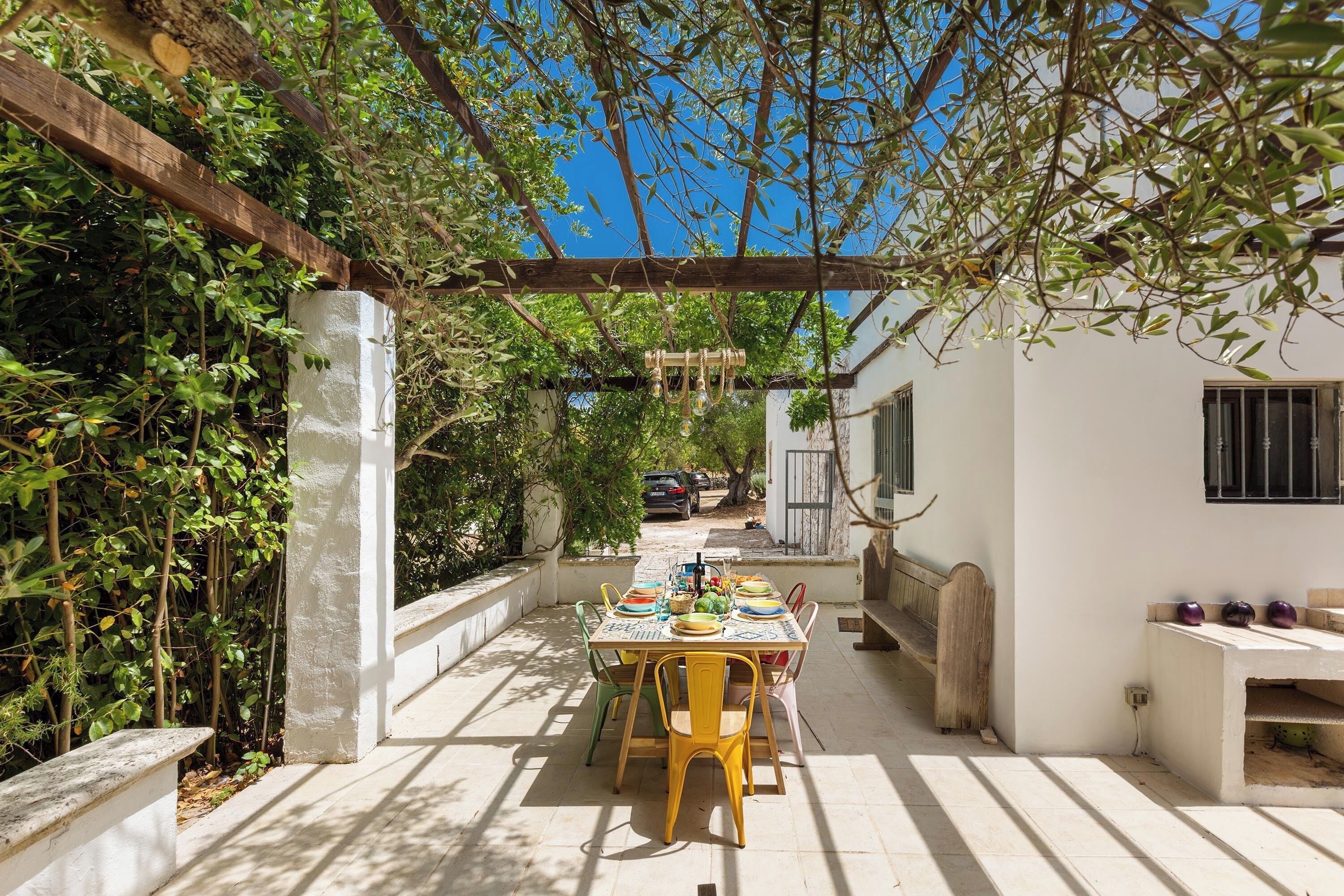 An outdoor dining area with a table set for four, surrounded by greenery and a pergola.