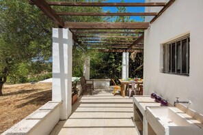 Uma área externa coberta com pérgola, uma mesa de jantar com cadeiras e vista para a paisagem ao redor.