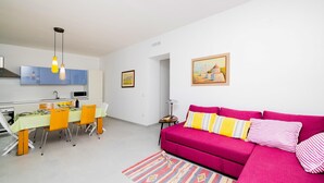 Un salon moderne avec un canapé rose vif, une table à manger avec des chaises et un coin cuisine doté de meubles bleus et d’un luminaire suspendu.