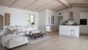 A spacious living room with a sofa, a coffee table, and a wicker chair. A kitchen area is visible with a countertop and a stool. - Stunning Ocean View and Private Beach (Nykobing Sjaelland)